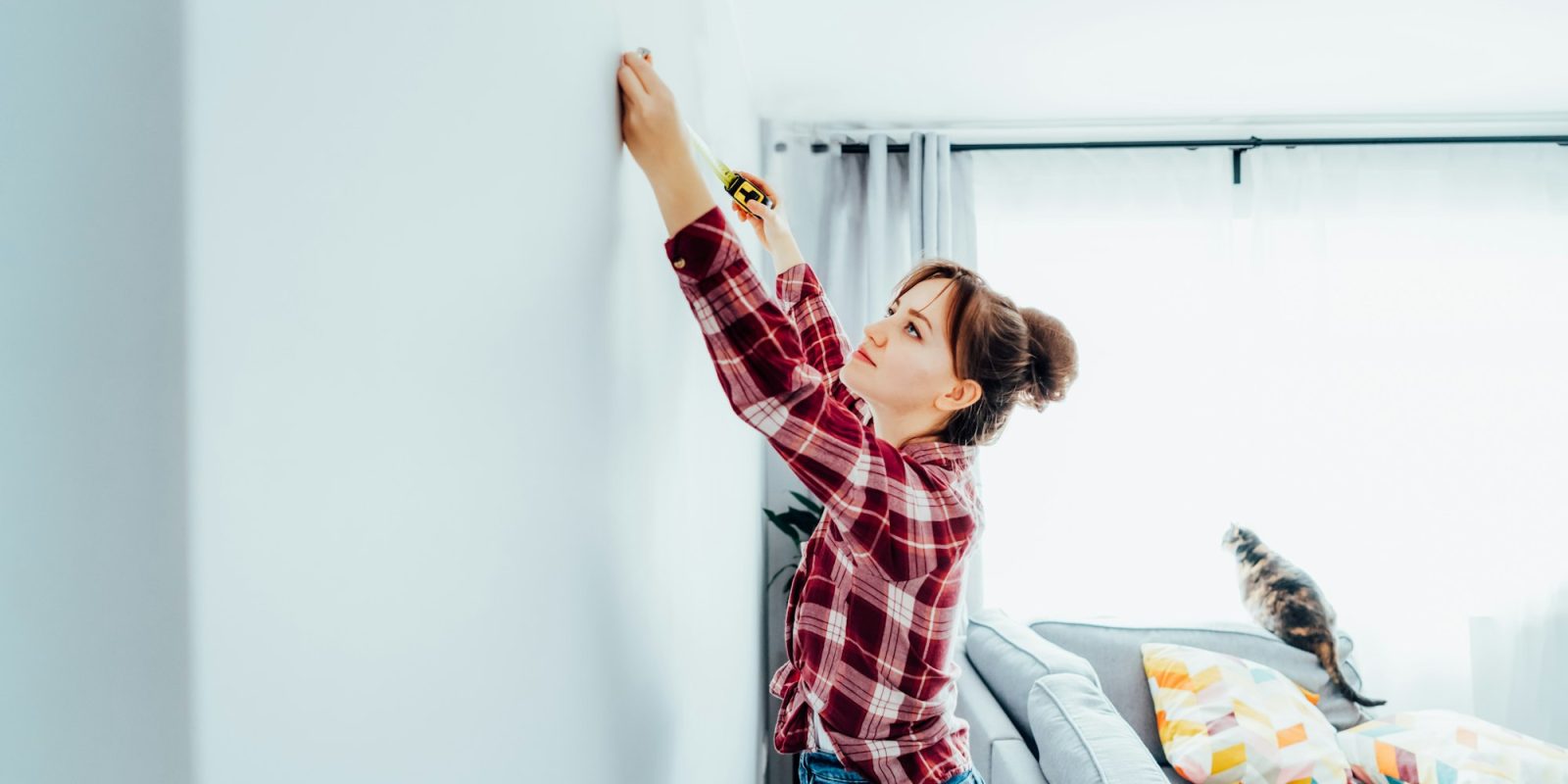 Young woman in plaid shirt doing measuring with measure tape on the wall. Doing repair herself. DIY