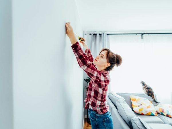 Young woman in plaid shirt doing measuring with measure tape on the wall. Doing repair herself. DIY