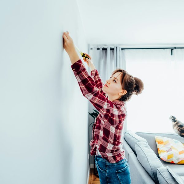 Young woman in plaid shirt doing measuring with measure tape on the wall. Doing repair herself. DIY