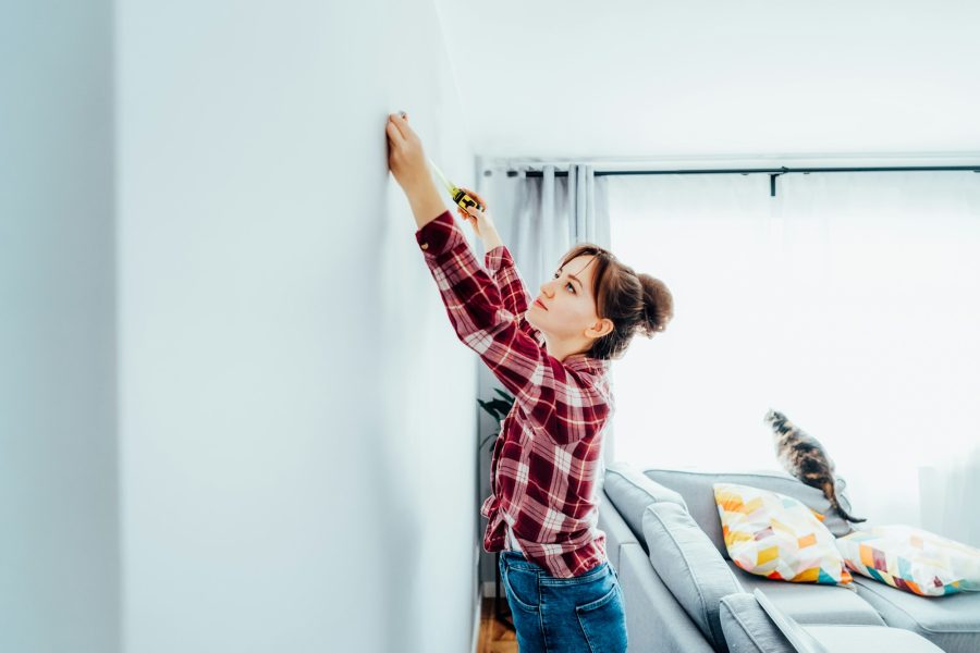 Young woman in plaid shirt doing measuring with measure tape on the wall. Doing repair herself. DIY